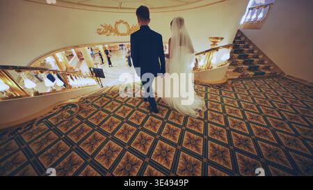 Elegant newlyweds gracefully walking down grand staircase, celebrating matrimonial union in opulent wedding venue Stock Photo