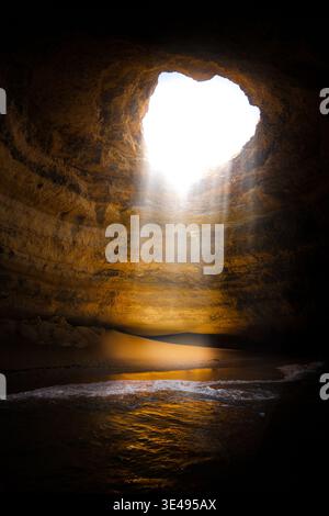 View of the sunlight streaming through the ceiling of Benagil Cave onto the sandy beach and ocean waves in Portimao, Faro District, Portugal. Stock Photo