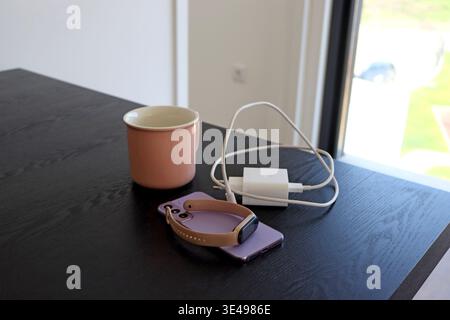 Smartphone with smart watch, charger cable and ceramic mug placed on dark table. Everyday technology setup for charging routine, digital lifestyle, co Stock Photo