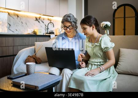 Senior woman with granddaughter using laptop while sitting together, browsing online content and sharing digital learning at home. Useful for family technology, online education, digital inclusion Stock Photo