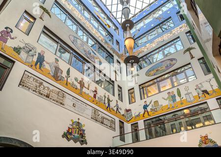 A panoramic view of the multi-storey atrium in the Speck's Hof exhibition hall, featuring surrounding mosaic friezes, a glass roof and a central pendant light; typical Leipzig exhibition hall architecture. Leipzig, Saxony, Germany Stock Photo