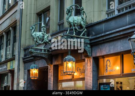 Two patinated bronze sculptures of boys on stone goats flank the entrance to an Art Nouveau commercial building; illuminated lanterns hang beneath the entrance arch. Leipzig, Saxony, Germany Stock Photo