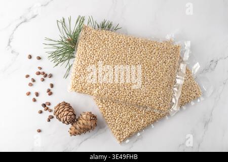 Vacuum packaging of shelled cedar pine nuts with cones and evergreen branch on marble background. Top view supermarket product in transparent package. Stock Photo