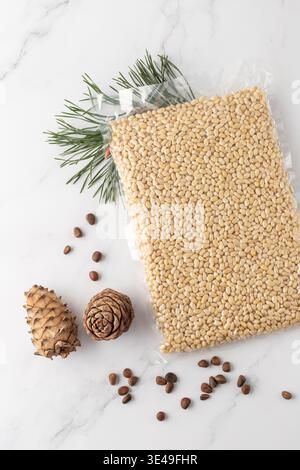 Vacuum packaging of shelled cedar pine nuts with cones and evergreen branch on marble background. Top view supermarket product in transparent package. Stock Photo
