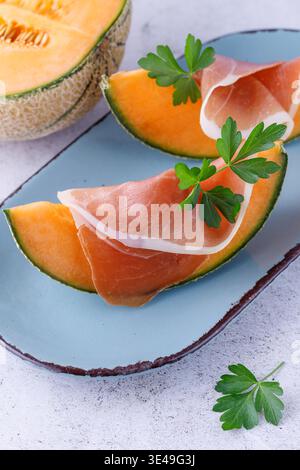 Traditional spanish jamon slices (dried pork meat) with melon (cucumis melo) and fresh parsley leaves herbs on vintage plate. Dish on light stone tabl Stock Photo