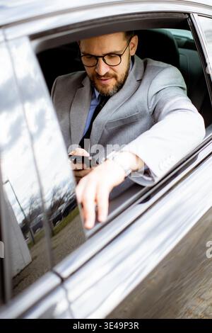 A businessman is seated in a black car. He is wearing glasses and checking his phone while holding it in his right hand. The car is parked in an urban Stock Photo