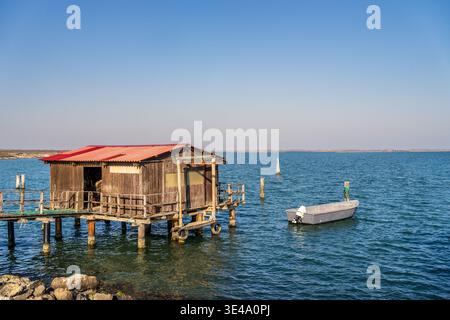 A characteristic wooden fisherman's hut on stilts with a red roof overlooks the calm blue waters of Sacca degli Scardovari. A small grey boat is moore Stock Photo