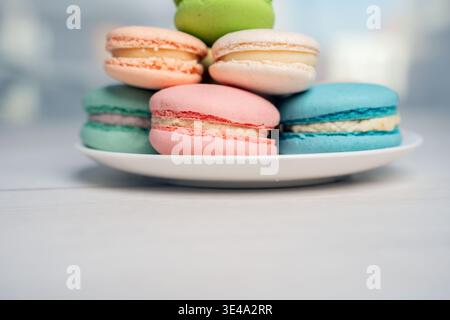 close-up of pastel macarons stacked on a white plate on a pale wooden tabletop, soft airy high-key composition showcasing pink, blue, green and cream Stock Photo