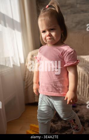 toddler in a pink shirt standing by a sunlit window with a furrowed, pensive expression; soft beige living room textures, warm natural light, scattere Stock Photo