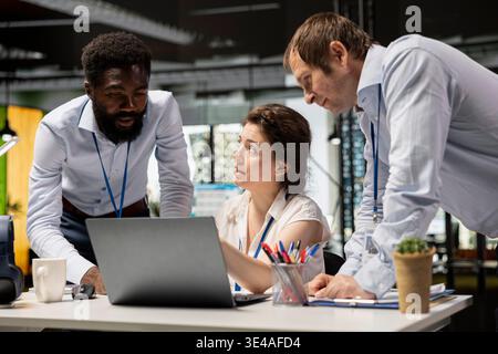 Diverse corporate employees in office workspace review financial data and investment reports on laptops. Professional woman conducts project briefing with male colleagues, analyzing company statistics Stock Photo