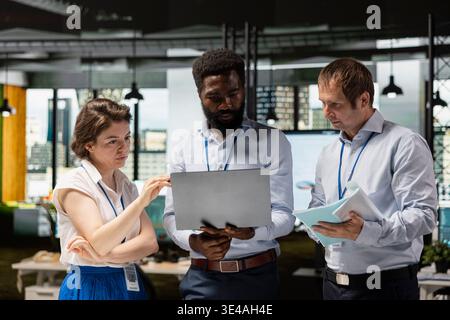 Black man and two white colleagues working together on financial reports and business investment strategies. Multiethnic office team standing and reviewing company performance on laptop. Stock Photo