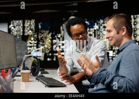 Upbeat programmers laughing, sharing jokes in startup office while optimizing code for performance and scalability. Jolly coworkers talking while using programming languages on computer Stock Photo