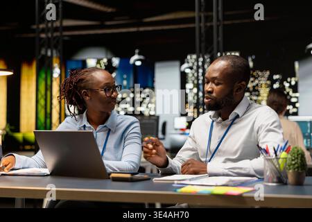 Helping young intern, laptop and manager in office completing spreadsheets, analyzing data. Assisting trainee, notebook and team leader, researching information online, working overnight Stock Photo