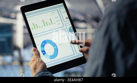 Close up of data center employee checking infrastructure diagnostics graphs on tablet screen. IT specialist on industrial platform in server room examining data charts during system audit, camera B Stock Photo