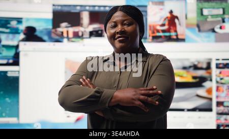 Portrait of cheerful woman celebrating freedom of unlimited video content in metaverse. Happy person stands with crossed arms in front of giant digital wall with online video channels, camera A Stock Photo