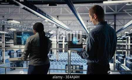 Data center system administrators investigate neural networks configuration, adjusting for optimal function. Server hub employees use laptop to check deep learning error reports, camera A Stock Photo