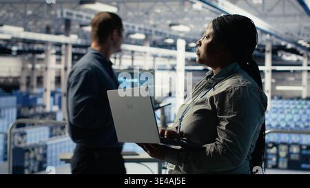Data center admin monitors deep learning process with laptop, reviewing operational metrics. African american woman using device to review neural networks reports during inspection, camera B Stock Photo