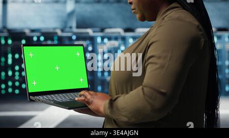 Programmer in data center using app on isolated screen laptop to ensure error free mainframes networks. Worker uses chroma key notebook to monitor server hub gear parts for failures, camera B Stock Photo