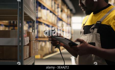 African american man using scanner to register barcodes tags in inventory, solving logistics operations in industrial warehouse. Worker checking shipping labels near storage racks. Camera B. Stock Photo