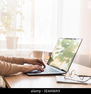 Womens hands typing on laptop keyboard, working remotely from home office desk with mug and glasses. Person typing on laptop working from home Stock Photo