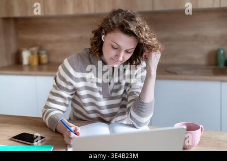 Enthusiastic woman student studying at home writing notes, listening online webinar wearing earbuds. Stock Photo