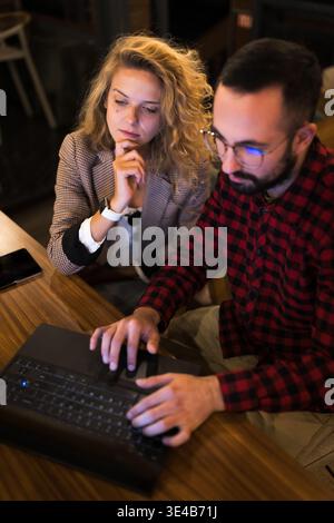 Two colleagues engage in a thoughtful discussion while working on a laptop in a cozy, dimly lit cafe setting, highlighting collaboration and teamwork Stock Photo
