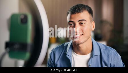 Happy man, influencer and recording with ring light in house for content creation, smile and video on phone. Person, web chat and live streaming with Stock Photo