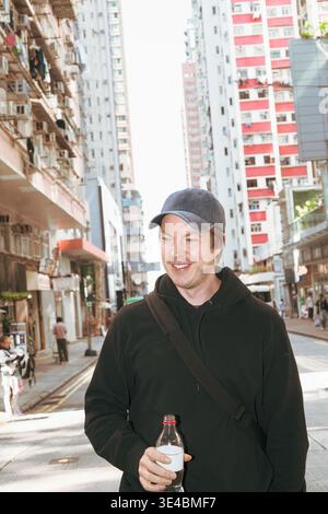 A man in a black hoodie and baseball cap smiles while holding a bottle on a sunny urban street in Hong Kong. High-rise apartment buildings fill the ba Stock Photo