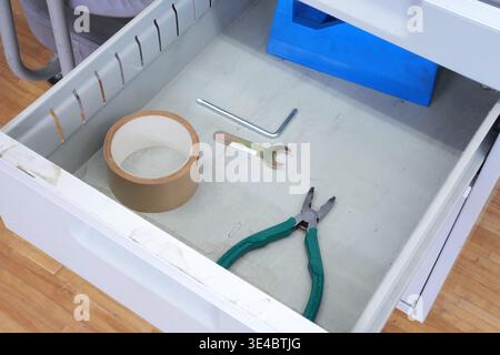 White drawer holds tools neatly arranged. Roll of tape, wrench, pliers, and Allen key rest inside. Blue plastic organizer sits tucked in the back. Cle Stock Photo