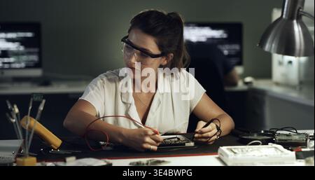 Soldering, woman or motherboard repair in office for hardware diagnostics, continuity or safety. Night, technician and multimeter probe on circuit for Stock Photo