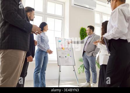 Diverse group of professionals in modern office, leader pointing at flip chart with diagrams, ideas Stock Photo