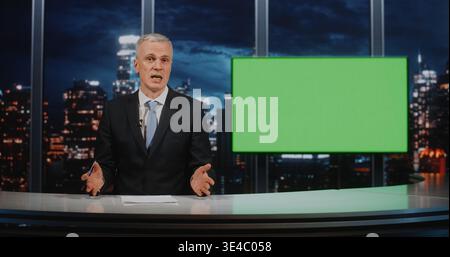 Male Anchor in Suit Sits at Desk With Papers and Pen. Near Man Large Green Chroma Key Screen, Ready For Virtual Background Insertion. Concept of Television Production and News Broadcasting Preparation Stock Photo