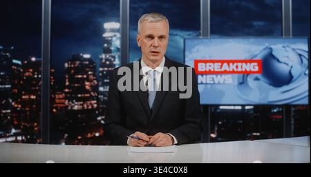 Serious Grey Haired Man Journalist in Blue Tie Sitting at White Desk With Papers, Presenting Information. Concept of Media Broadcasting, Professional Portrait Against Blurred Urban Nighttime Vista. Stock Photo