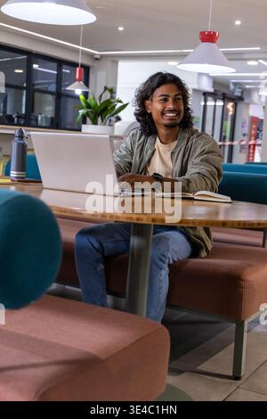 Indian man mid-20s smiling while working on laptop at workspace with notebook, bomber jacket Stock Photo