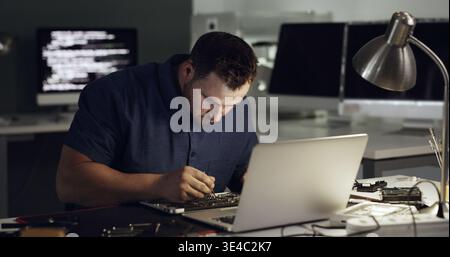 Technician, night and man on laptop for repair, maintenance and troubleshooting with hardware. Office, IT and person on computer with tools for Stock Photo