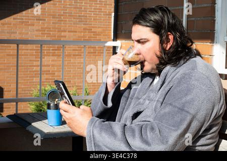Man wearing a bathrobe drinking espresso and looking at smartphone on a balcony Stock Photo