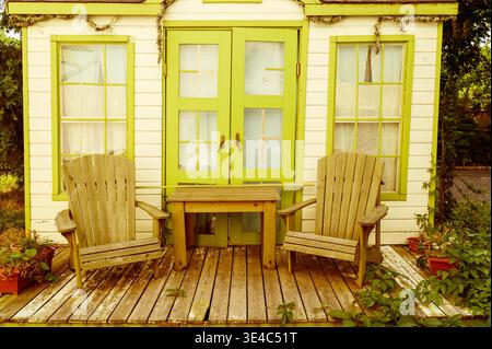 Rustic wooden decking area with two armchairs and a table near a warm colored house wall. Blooming flowers. Concept of cozy country hospitality, aesth Stock Photo