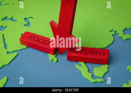 A red downward arrow breaks apart wooden blocks reading FOOD and SECURITY over a world map. Global food crises, agricultural supply chain disruptions Stock Photo
