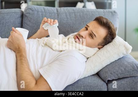 Sick man with runny nose resting on sofa at home, holding tissue and hot drink Stock Photo