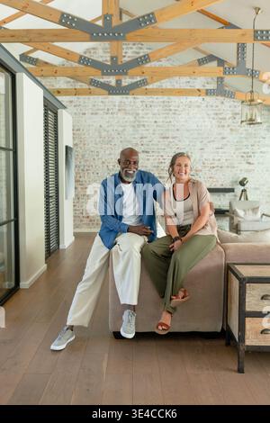 Diverse couple sitting on beige sectional sofa, leaning together in living room with brick wall Stock Photo