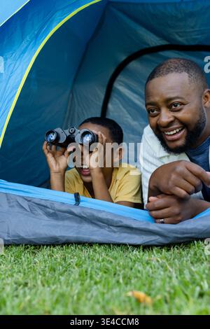 African American father and son lying at pop-up tent on lawn, boy holding black binoculars Stock Photo