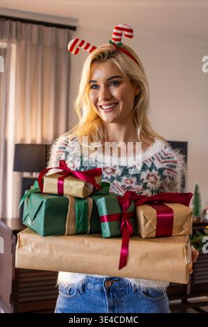 Woman in her 20s holding wrapped gifts at home, wearing fuzzy sweater and candy cane headband Stock Photo