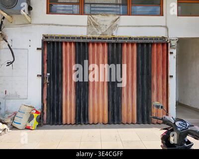 A closed vertically ribbed folding metal security gate with alternating black and rust-colored panels, installed on a building facade. Indonesia, Bata Stock Photo