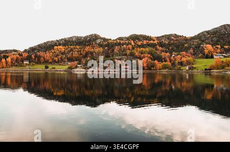 A tranquil coastal landscape featuring colorful deciduous trees and small seaside cabins reflected perfectly in calm water beneath rugged mountain slo Stock Photo