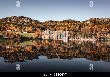A tranquil autumn landscape featuring colorful deciduous trees and small lakeside cabins reflected perfectly in calm water beneath rugged mountain slo Stock Photo