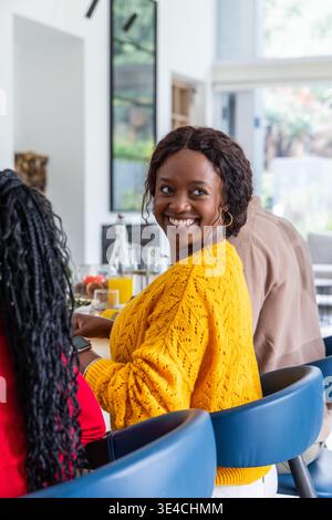 Diverse friends in kitchen, woman in yellow sweater smiling on blue bar stool, chatting at counter Stock Photo