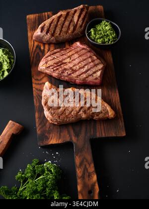 Three pieces of grilled roast beef flank and rump steaks are placed on a wooden board. Green sauce is served in small bowls alongside. Fresh parsley i Stock Photo