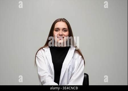 Young woman confidently smiling, wearing a white shirt, sitting relaxed against a plain background in a studio Stock Photo