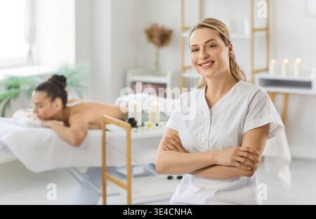 Portrait of smiling female massage therapist in white uniform posing in spa room with client behind. Stock Photo