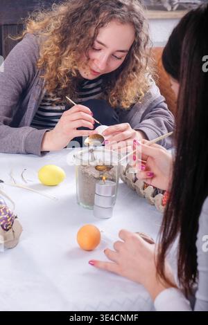 Young women decorating easter eggs with wax technique at table focused hands painting patterns beside candle and tools creating creative festive indoo Stock Photo